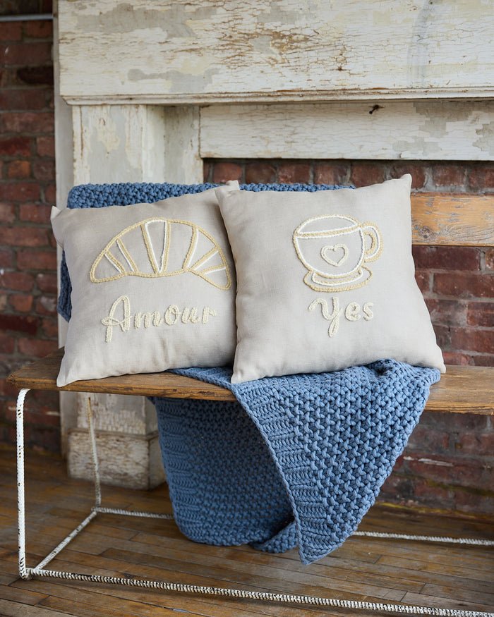 Beige square pillow with an embroidered coffee cup and the word 'yes'. Beige square pillow with embroidered croissant and the word 'amour'.