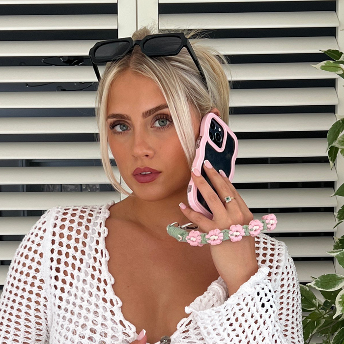 Woman in sunglasses and white crochet top holds a pink phone with a crochet flower phone strap bundle attached.