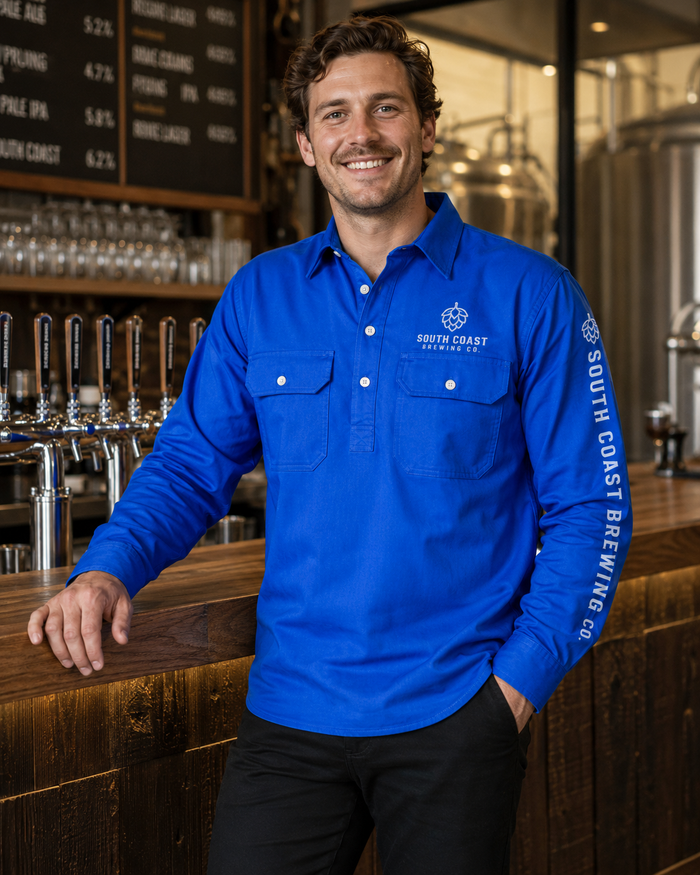 A smiling man stands behind a wooden bar counter wearing a bright blue long-sleeve button-up shirt with white South Coast Brewing Co. logos on the chest and sleeve, paired with black pants in a brewery setting.