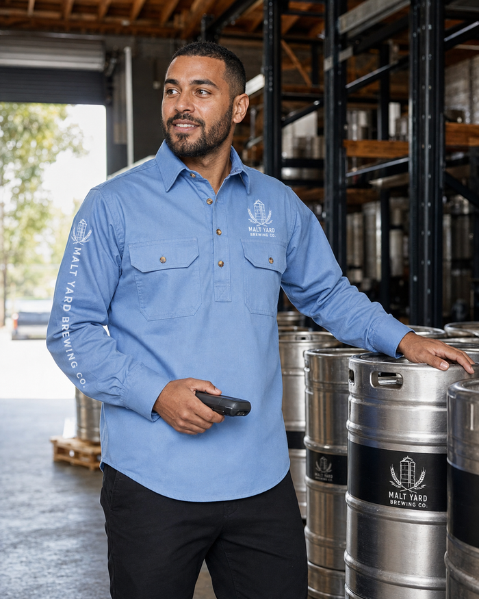 A man wearing a light blue long-sleeve button-up shirt with a printed logo stands in a brewery warehouse next to stainless steel beer barrels.