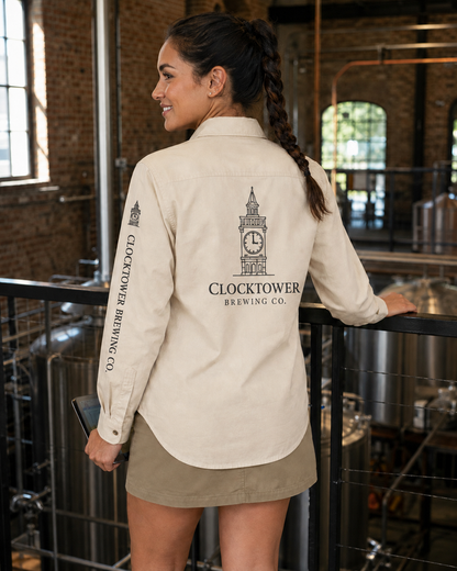 A woman stands in a brewery wearing a cream-colored long-sleeve button-up shirt with a black printed Clocktower Brewing Co. logo on the back and sleeve, paired with a khaki mini skirt.