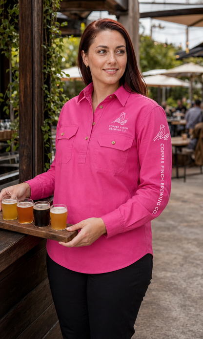 A woman in a bright pink long-sleeve button-up shirt stands at an outdoor bar holding a wooden tray with four beer glasses. The shirt has white printed logos on the chest and sleeve, including 'Copper Ridge Brewing Co.