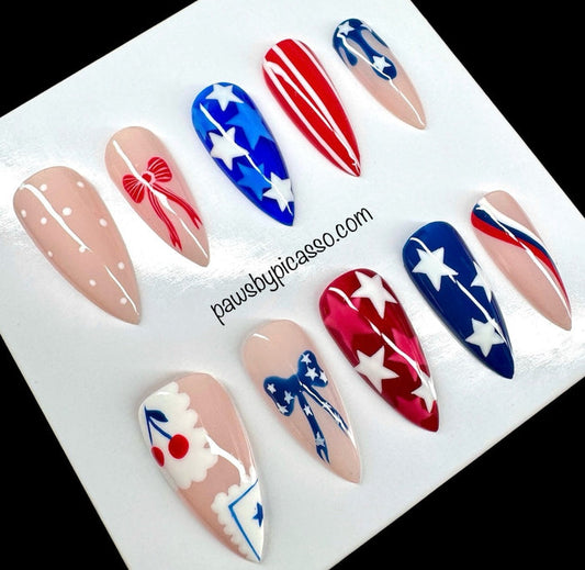 Ten press-on nails with patriotic red, white, and blue designs, including stars, stripes, and bows, displayed on a white card with the text 'pawsbypicaso.com'.