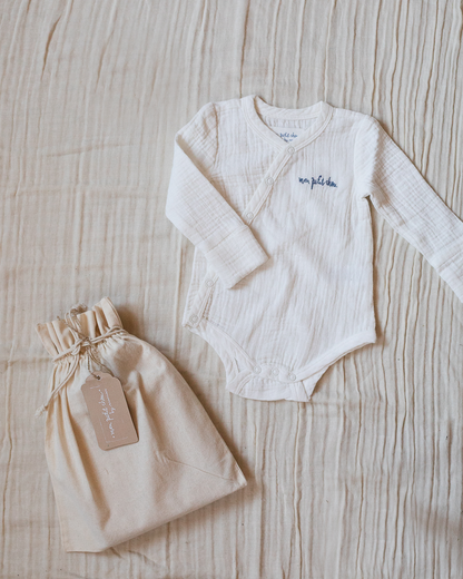 White muslin bodysuit with "mon petit chou" ("my little darling" in French) embroidered in blue on the chest, placed next to a small beige toile bag.